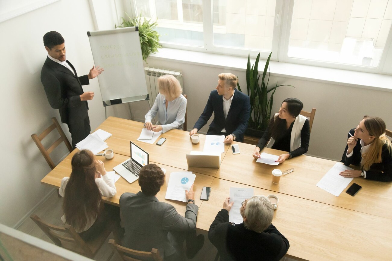 Personen an Tisch sitzend hören Vortrag eines Referenten zu, der vor Whiteboard steht